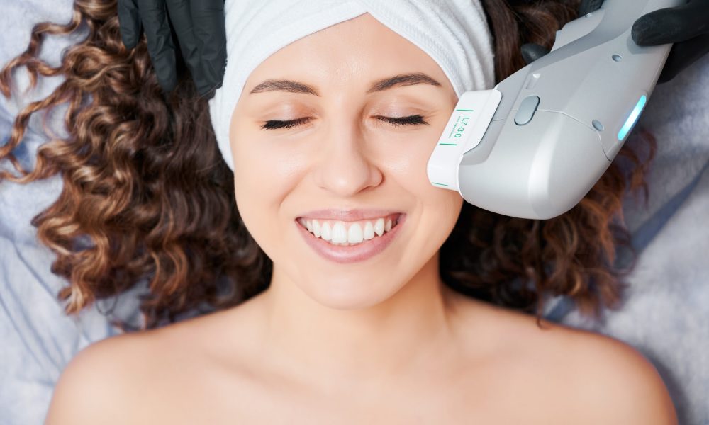 Cheerful young woman having face lifting procedure in clinic. Top view of beautiful young woman with closed eyes wearing white headband while receiving facial treatment in cosmetology clinic. Charming female patient having face lifting procedure and smiling.